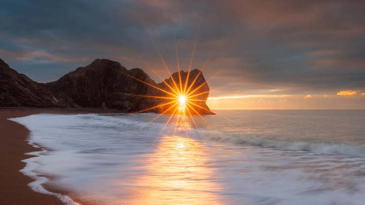Image "The Moment" - seashore scene with sunrise shining through a gap in the rocks